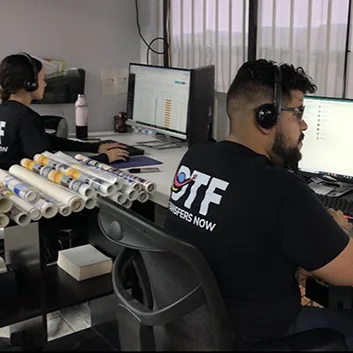 Two people wearing headsets work at desks with computers. Several rolls of printed material are stacked next to them. They are wearing matching black shirts with a logo and text. The room has light purple walls and vertical blinds.